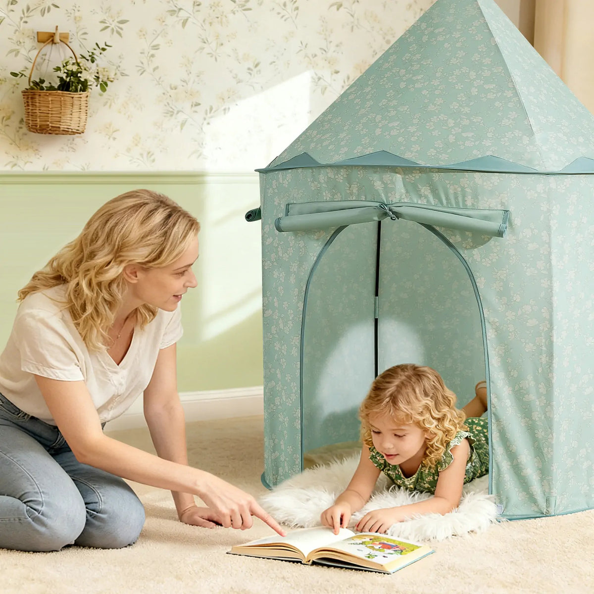A woman kneels on the floor pointing to a book for a young child lying inside a pale green floral Starry Indoor Play Tent for Toddlers.