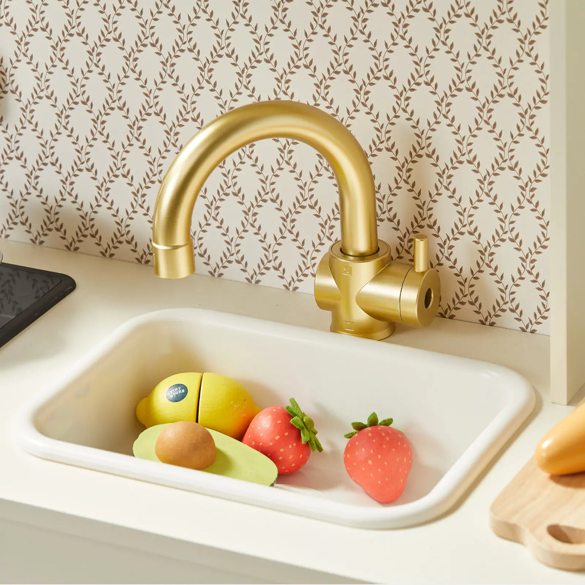 White PopJoy Play Kitchen with Lights & Sounds sink featuring a gold faucet and toy fruit, including a lemon, strawberries, and an avocado.