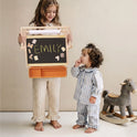 A child holds a 3-in-1 Adjustable Tabletop Art Easel with a chalkboard spelling EMILY in letter stickers, with a younger child standing beside it.