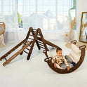A young child sits at the base of a Chocolate 5in1 Montessori Climbing Set—featuring a Triangle Ladder, Arch/Rocker, Slide Board/Ramp, Netting Rope, and Cushion—surrounded by light pillows in a bright, cozy room with large windows.