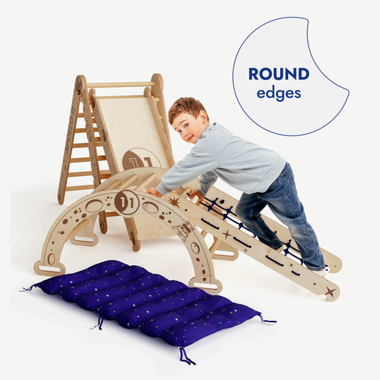 A young boy climbs the Apollo 5in1 Climbing Set: Pikler Triangle, Net, Arch, Slide Board, and Cushion for Little Astronauts. The image shows the blue padded mat on the floor and a ROUND edges label. The set is made of light wood.