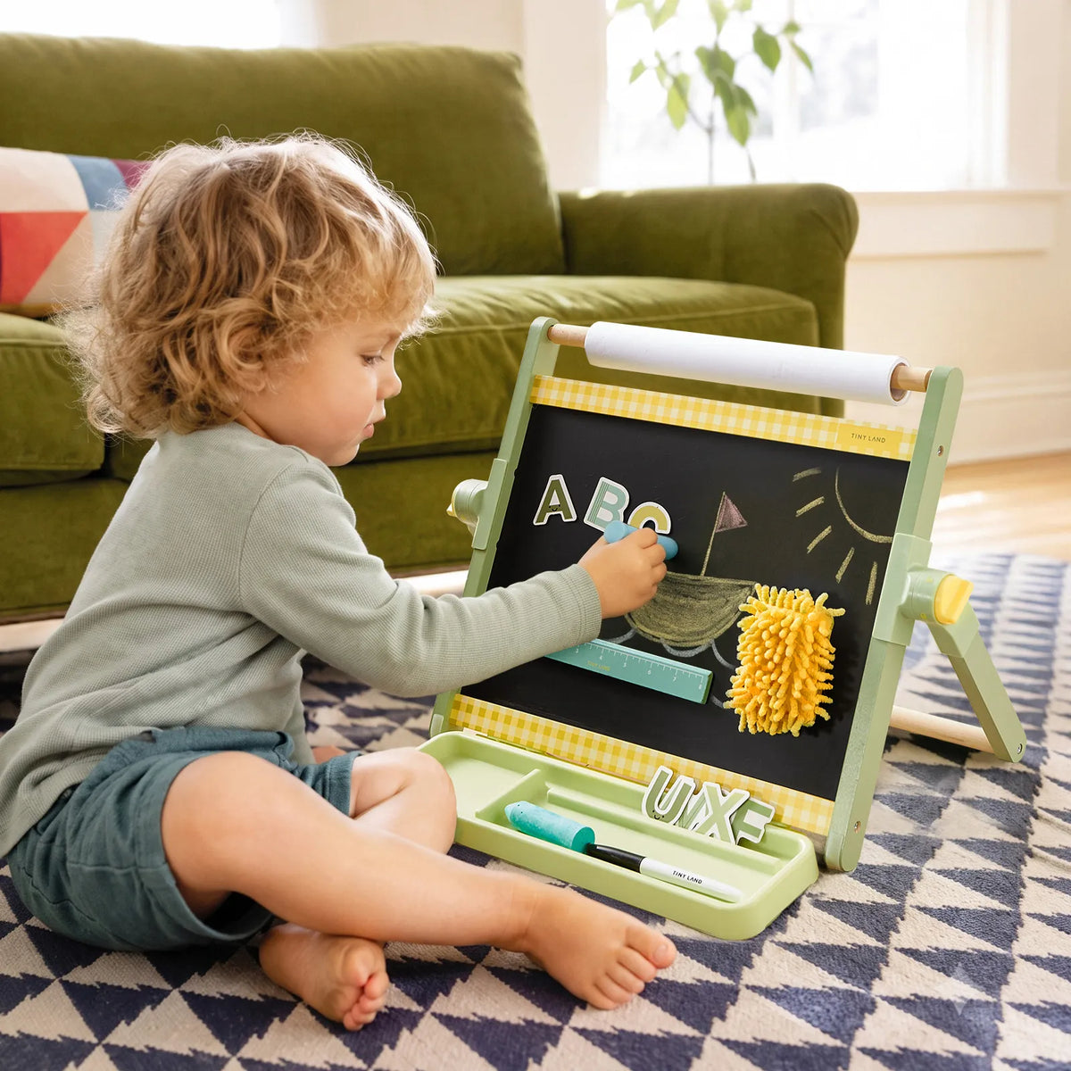 A toddler sits on a rug playing with a 3-in-1 Adjustable Tabletop Art Easel that features a chalkboard with magnetic letters, a drawing of a sailboat, and a paper roll holder.