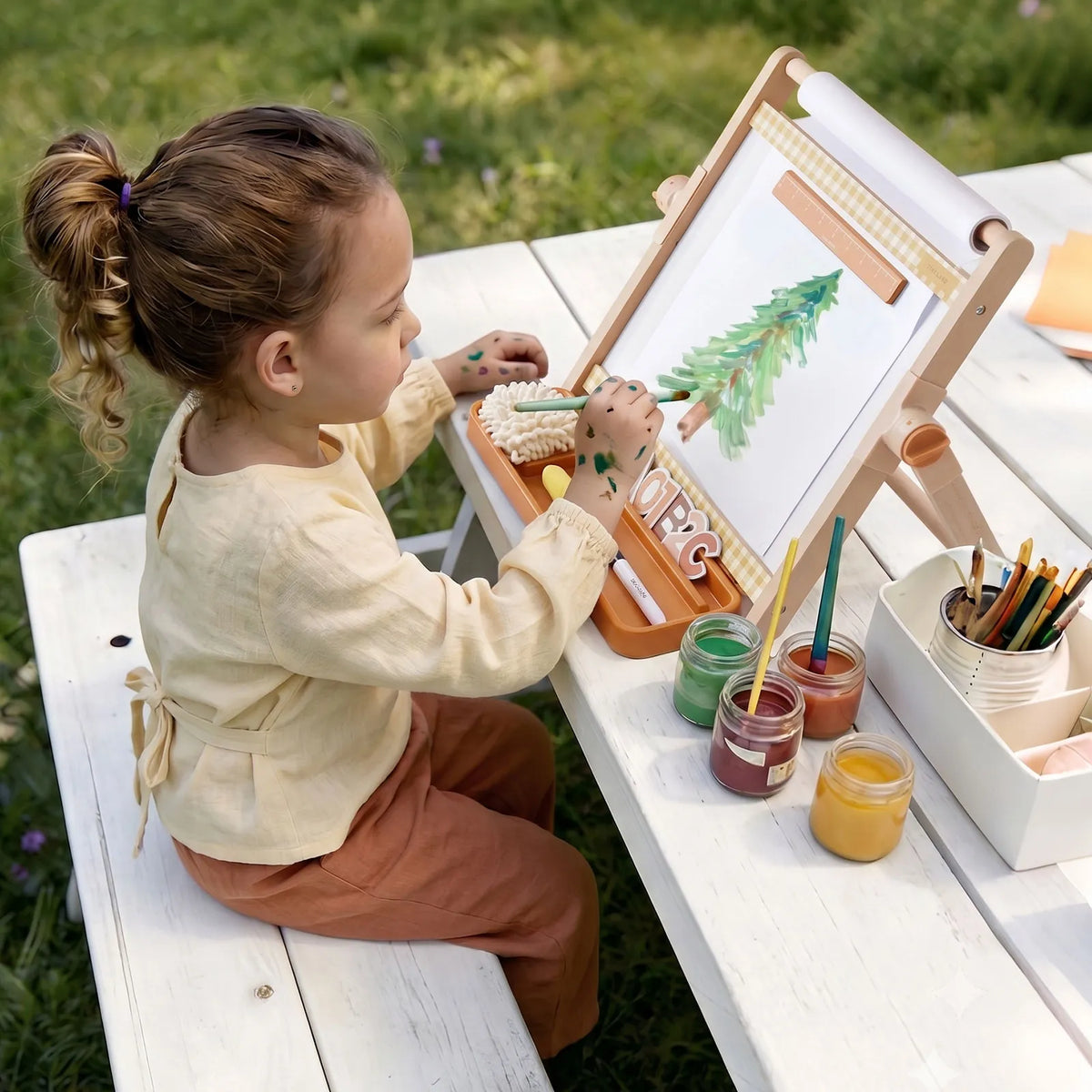 A young child paints a picture of a pine tree on a 3-in-1 Adjustable Tabletop Art Easel set up on a white outdoor bench.