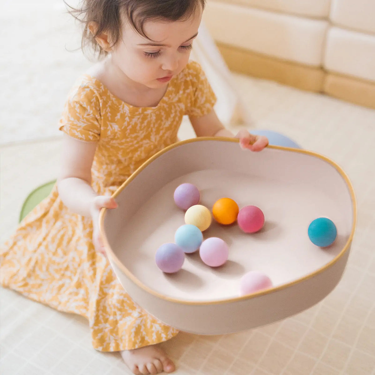 A toddler holds a shallow bowl from a Stacking Balance Stepping Stones – Sunset set, which contains several small, colorful spheres.