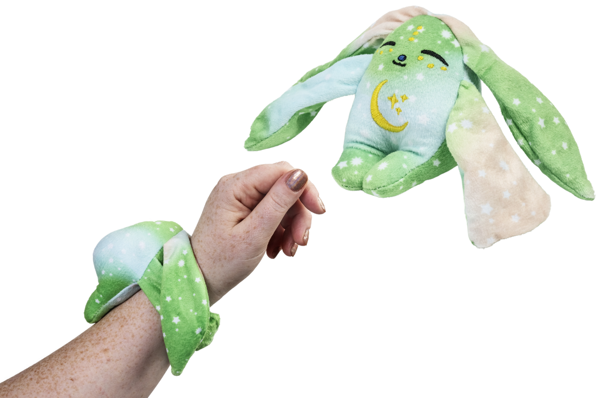 A person wears a green Clover Mini Wrist Hugger – Magnetic Sensory Plush while a matching bunny toy floats in the air nearby.