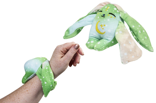 A person wears a green Clover Mini Wrist Hugger – Magnetic Sensory Plush while a matching bunny toy floats in the air nearby.