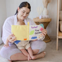A woman sits cross-legged, smiling as she reads Tiny Land® Montessori Toys for Newborns (4–6 Months)—a colorful soft book—to a baby on her lap, creating a cozy moment and offering one of the perfect sensory toys for babies.