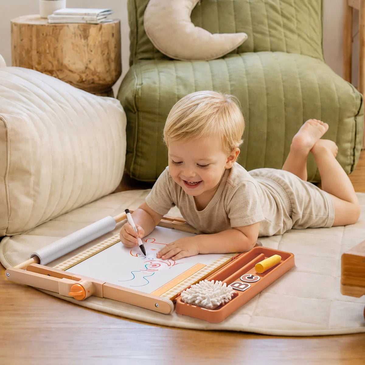 A young child lies on a play mat drawing on a 3-in-1 Adjustable Tabletop Art Easel, which features an attached tray of chalk and magnetic letters.