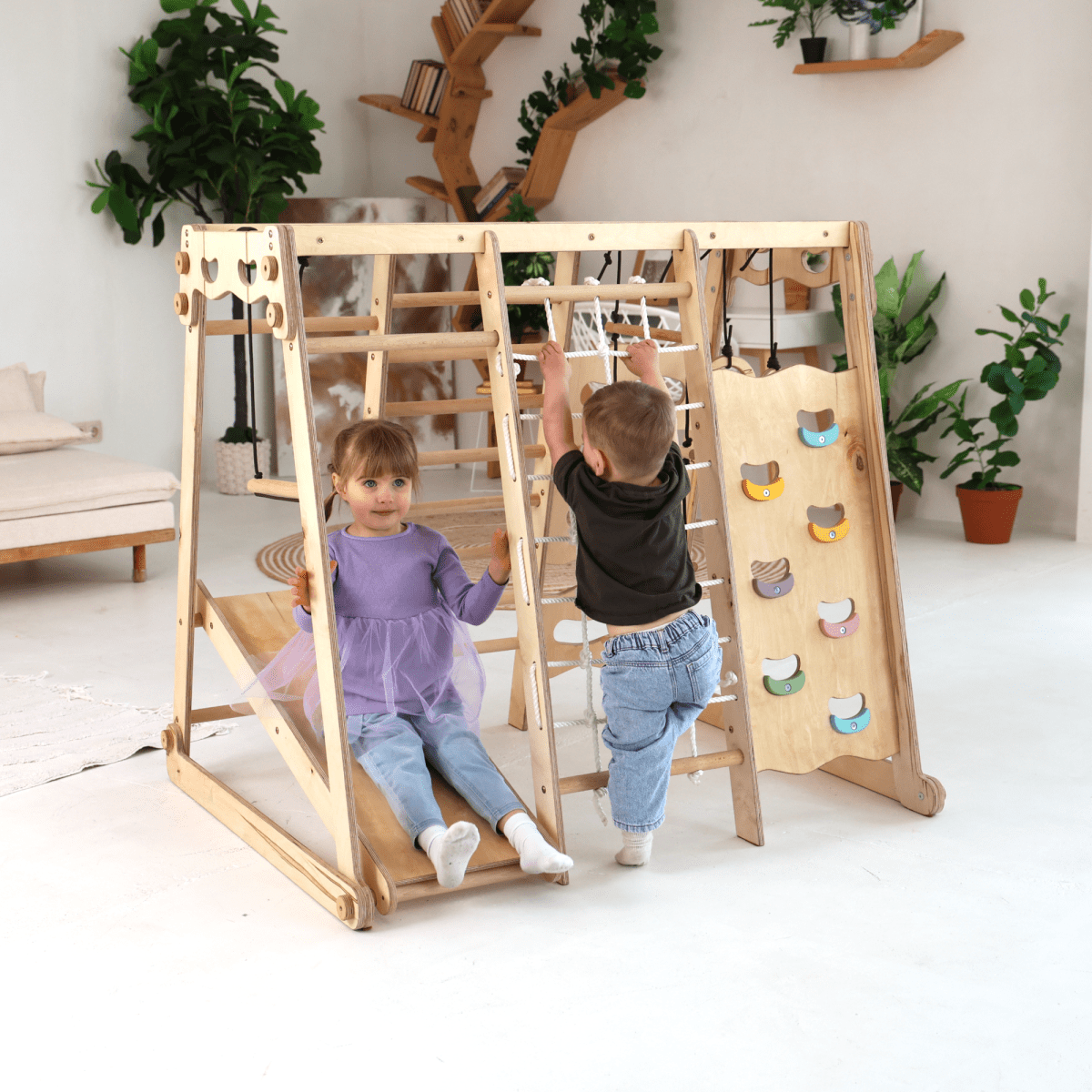 Two young kids play on the 6in1 Indoor Jungle Gym for Toddler, featuring a playground, swings set, slide board, rope, and stairs. The girl in a purple dress sits on the slide; the boy in black scales the rope net in a bright room with green plants.