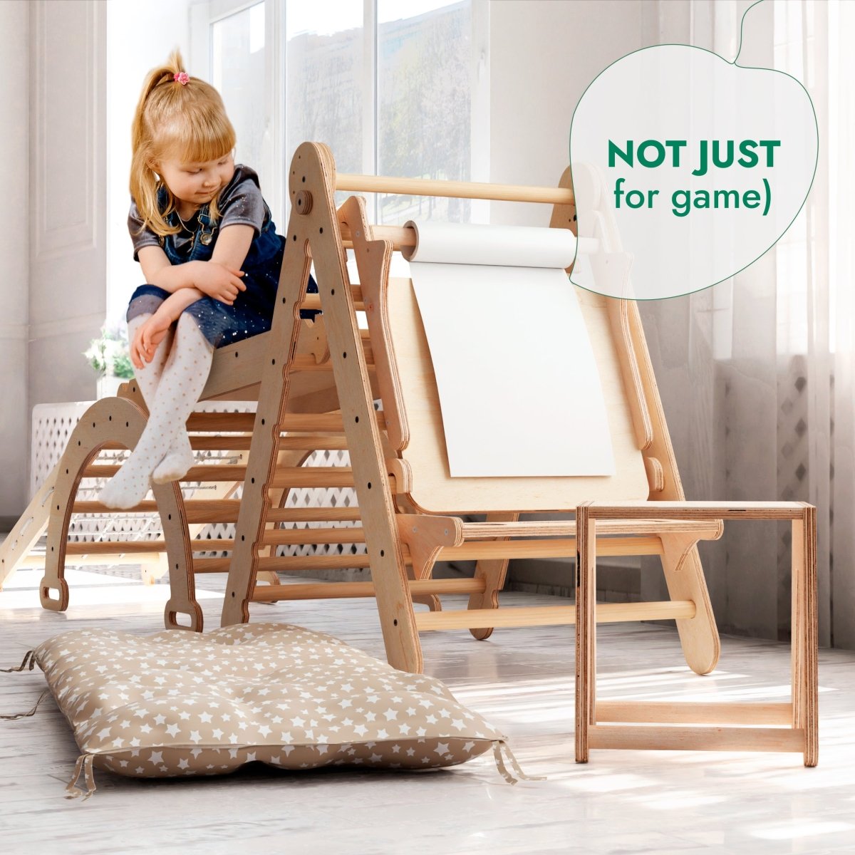 A young girl sits on the 6in1 Montessori Climbing Set: Triangle Ladder + Arch/Rocker + Slide/Ramp + Net + Cushion + Art Addition in a bright room with a cushion, small table, and a sign that reads “NOT JUST for game,” highlighting the multifunctional play equipment designed to support holistic child development.