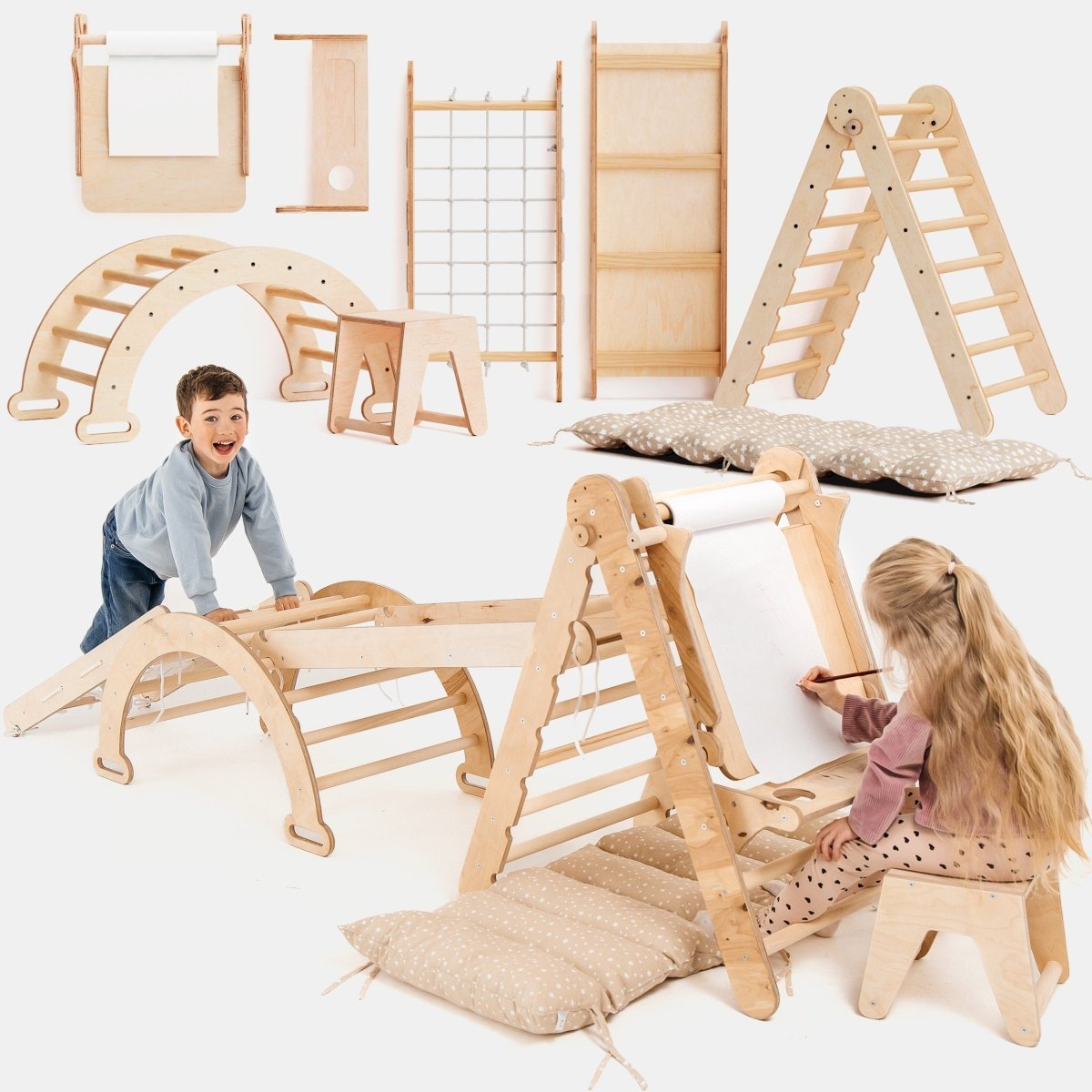 Two children play on the 6in1 Montessori Climbing Set, which includes a triangle ladder, arch/rocker, slide/ramp, net, cushion, and art addition. Cushions are placed on the floor for extra comfort. The light wood set is thoughtfully designed to support holistic child development through safe and engaging play.
