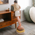 A child stands on a stack of four colorful, bowl-shaped Stacking Balance Stepping Stones – Sunset while reaching onto a wooden console table.