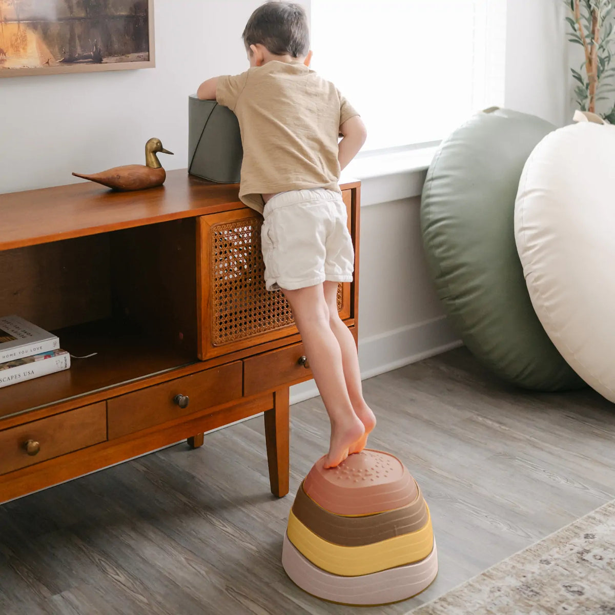 A child stands on a stack of four colorful, bowl-shaped Stacking Balance Stepping Stones – Sunset while reaching onto a wooden console table.