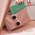 A hand with teal-painted nails places a green rabbit-illustrated book into a striped pocket of a Kids Beach Tent & Sand Toy Set, which features flower-shaped sunglasses attached to the front.