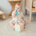 A baby in a beige knit romper sits on a carpet, pulling colorful tissues from the Tiny Land® Montessori Toys for Newborns (4–6 Months) sensory box, bringing one to their mouth. Wicker furniture appears in the background.
