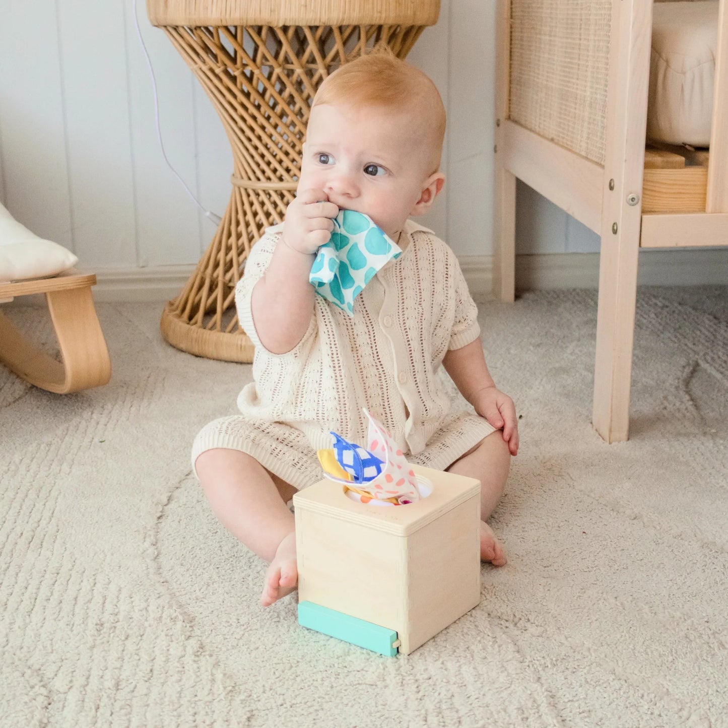 A baby in a beige knit romper sits on a carpet, pulling colorful tissues from the Tiny Land® Montessori Toys for Newborns (4–6 Months) sensory box, bringing one to their mouth. Wicker furniture appears in the background.