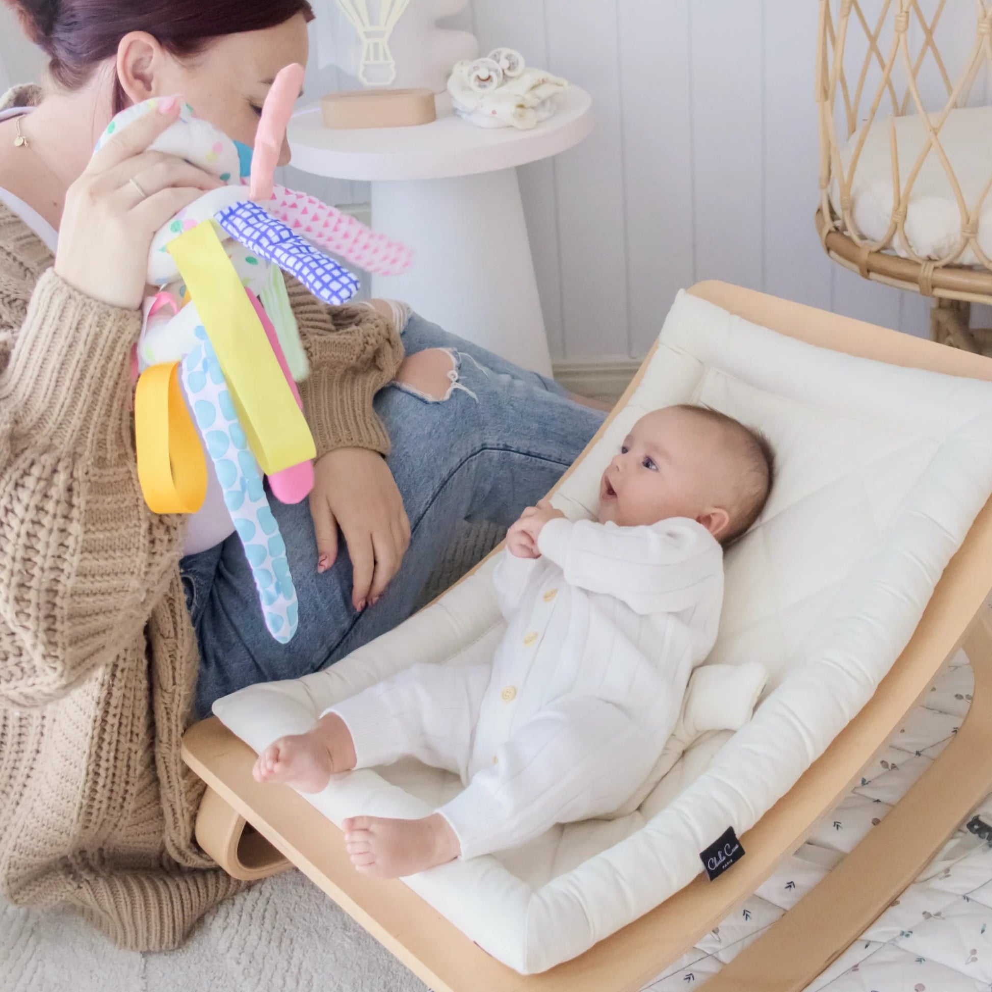 A baby in a white onesie lies in a cushioned rocker, gazing up at an adult holding the Tiny Land® Montessori Toys for Newborns (0–3 Months) set. The adult sits on the floor next to the rocker in a cozy, light-filled room.