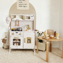A PopJoy Play Kitchen with Lights & Sounds featuring a stove, microwave, and sink, next to a small wooden table topped with pretend cooking food and a drinks menu sign.