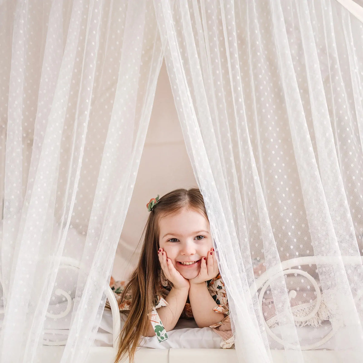 A young girl smiles with her chin in her hands, peeking through the white sheer curtains of a Large Cozy Cabin Play Tent with Star Lights.