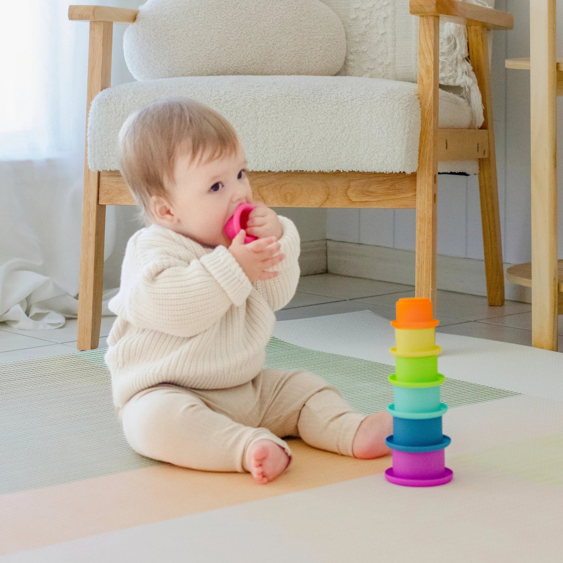 A baby in a cream-colored outfit sits on a mat, holding and mouthing a pink stacking cup from the Tiny Land® Montessori Toys for Newborns (7–9 Months), with other colorful stackable cups beside them and a wooden chair in the background.