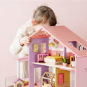 A young child leans in to play with a Sweetwood Cottage Dollhouse (48-pc Furniture), a multi-level wooden dollhouse furnished with miniature beds, a crib, and decor.