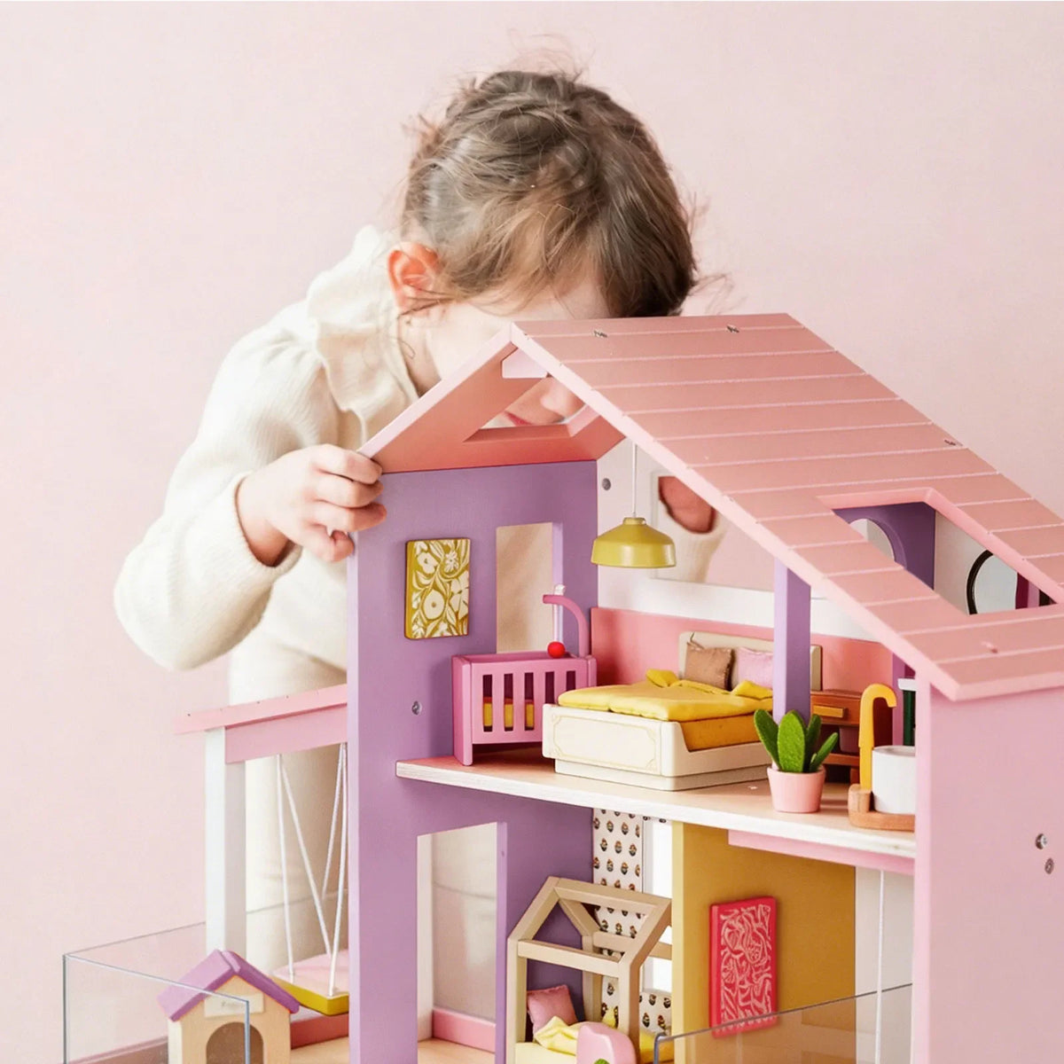 A young child leans in to play with a Sweetwood Cottage Dollhouse (48-pc Furniture), a multi-level wooden dollhouse furnished with miniature beds, a crib, and decor.