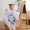 A young child smiles and holds up a colorful painting in front of the Tiny Land® Magnitales Easel for Kids, set in a bright, cozy room with a plant in the background.