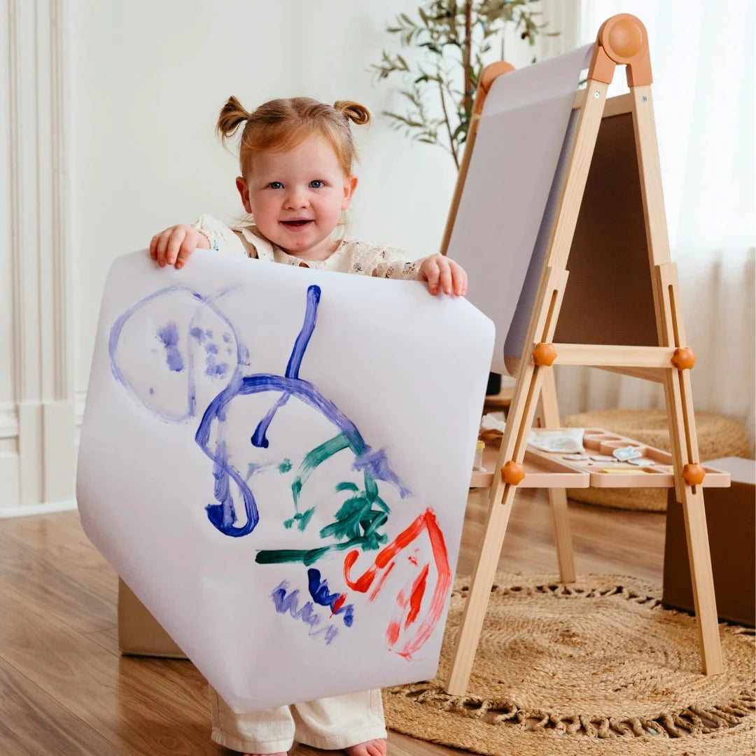 A young child smiles and holds up a colorful painting in front of the Tiny Land® Magnitales Easel for Kids, set in a bright, cozy room with a plant in the background.