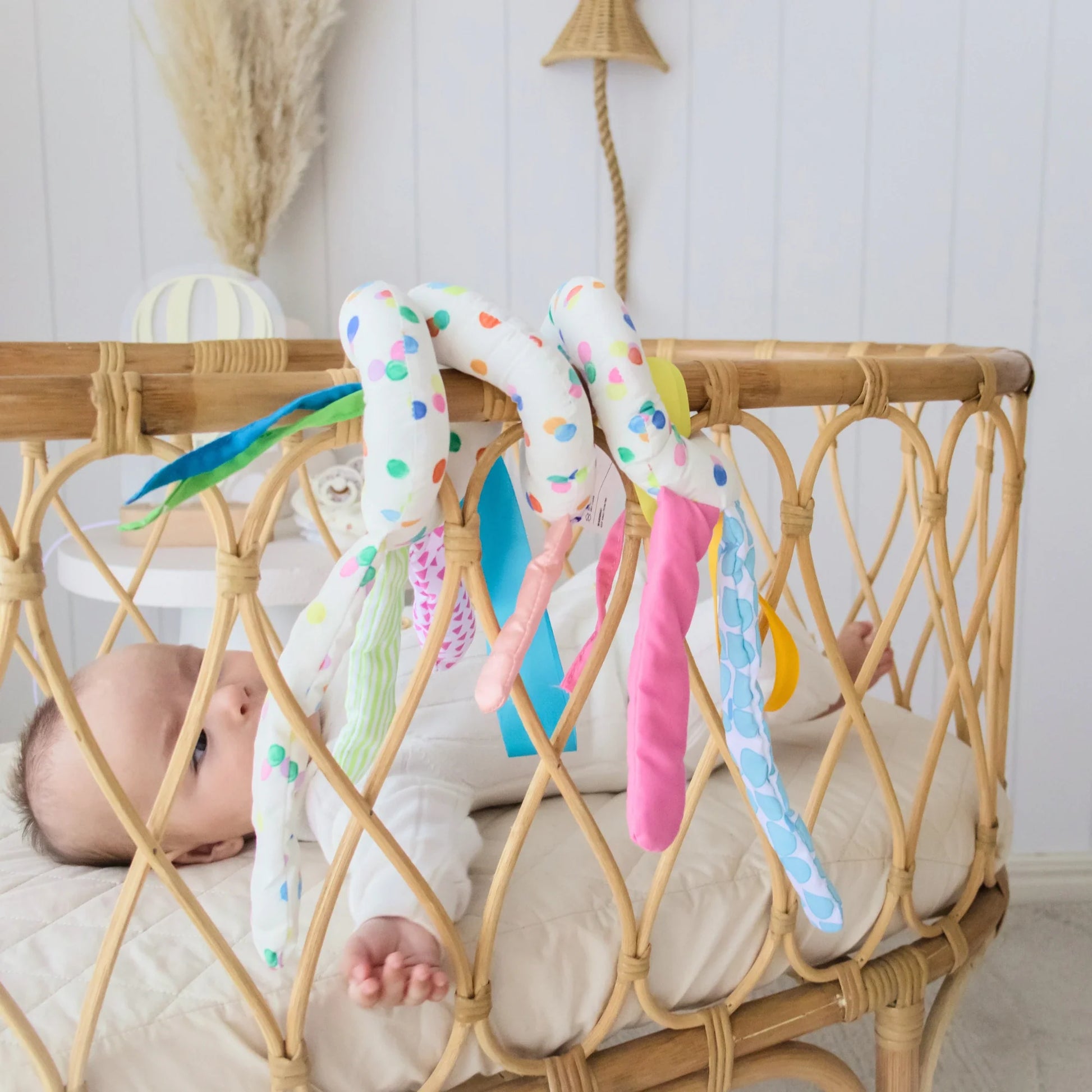 A baby lies in a wicker crib, reaching for Tiny Land® Montessori Toys for Newborns (0–3 Months). The light room with soft decor and pampas grass creates a soothing space, perfect for exploring these colorful sensory toys.