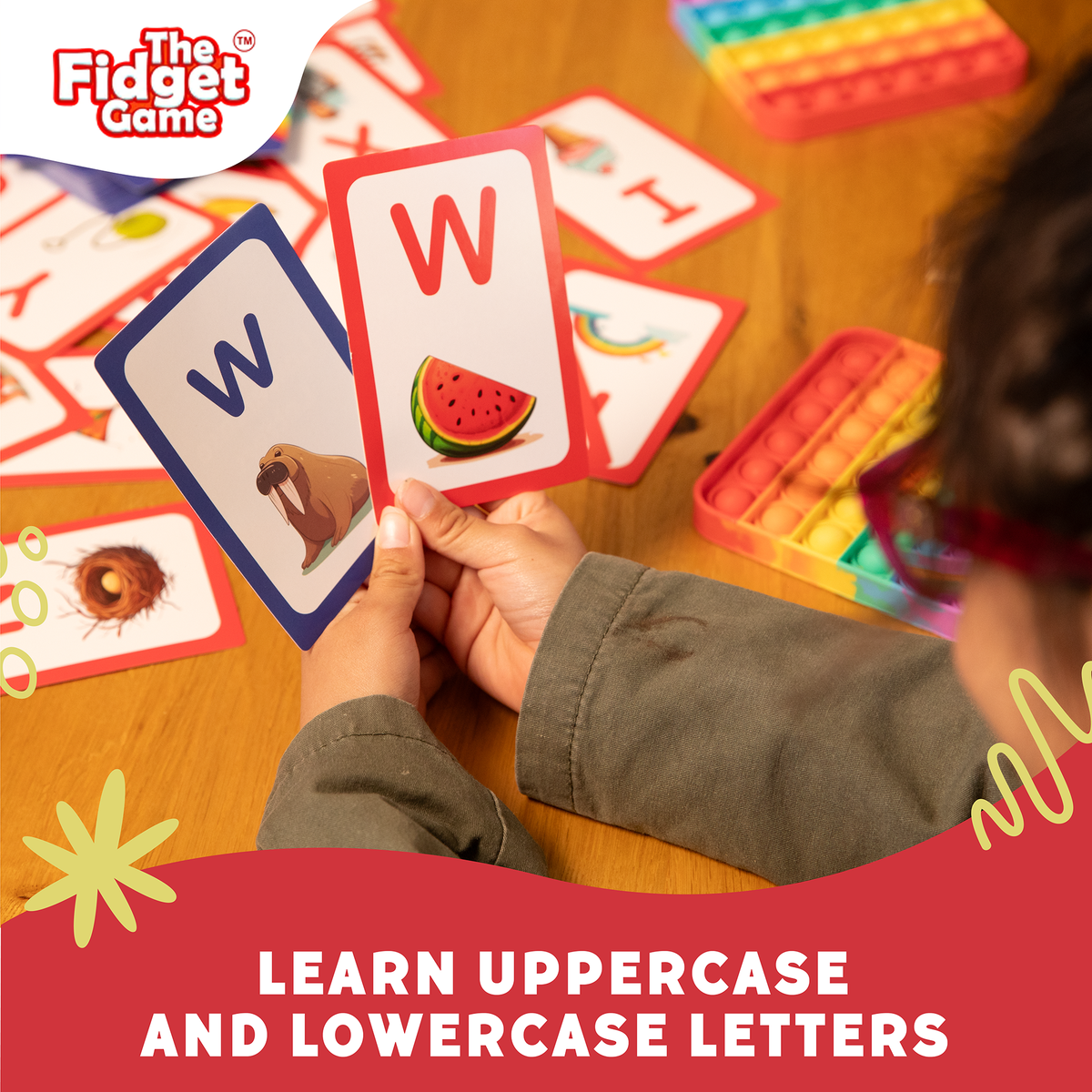 A person holds two ABC Uppercase & Lowercase Letters Card Pack flash cards showing the letter W, one with a walrus and the other with a watermelon, with a pile of cards and fidget toys on the table for letter recognition.