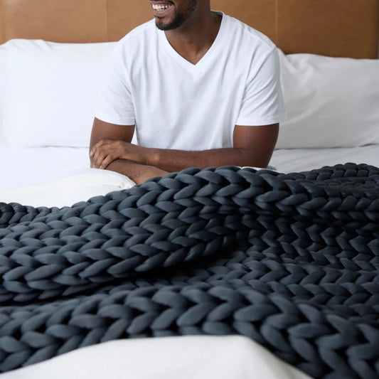 A man wearing a white t-shirt smiles while sitting on a bed, covered with the Ice Knit Weighted Blanket in thick, chunky dark gray.