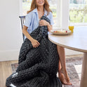 A woman smiles at a dining table with food and orange juice, wrapped in a chunky dark gray Ice Knit Weighted Blanket as sunlight streams through large windows in the background.