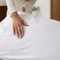 A person places their hand gently on a white sheet, possibly comforting someone or preparing a bed made with luxurious Forever Chill Bamboo Sheets, as soft natural light filters in the background.