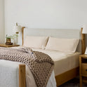 A neatly made bed features Forever Chill Bamboo Sheets in Tundra Beige, accented with a chunky knitted blanket. Wooden nightstands with lamps and a small plant flank each side. The room exudes a calm, minimalist style with neutral tones, elevated by the soft, cooling comfort of the bamboo viscose sheets.