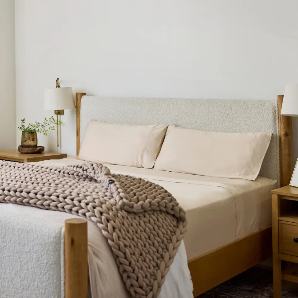 A neatly made bed features Forever Chill Bamboo Sheets in Tundra Beige, accented with a chunky knitted blanket. Wooden nightstands with lamps and a small plant flank each side. The room exudes a calm, minimalist style with neutral tones, elevated by the soft, cooling comfort of the bamboo viscose sheets.