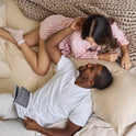 A couple relaxes on a bed dressed in Forever Chill Bamboo Sheets; the woman in striped pajamas gently touches the man’s face while he smiles at her, holding an open book. Cozy blankets and soft pillows crafted from organic bamboo viscose surround them, enhancing the serene atmosphere.