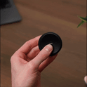 A hand presses the center of the ONO Slider Aluminum, a small, round calming fidget toy, making its outer ring glow blue. A laptop and a plant are visible blurred in the background.