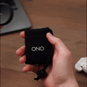 A hand holds a small black drawstring pouch labeled ONO in white letters, with a wooden surface, part of a laptop, and wireless earbuds in the background—ideal for carrying your ONO Slider Aluminum calming fidget toy.