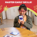 A young girl wearing pink glasses smiles while holding a counting flash card from the Card Pack Bundle (6 Learning Sets, 416 Cards) with a purple cube illustration, surrounded by other preschool math cards on a wooden table.