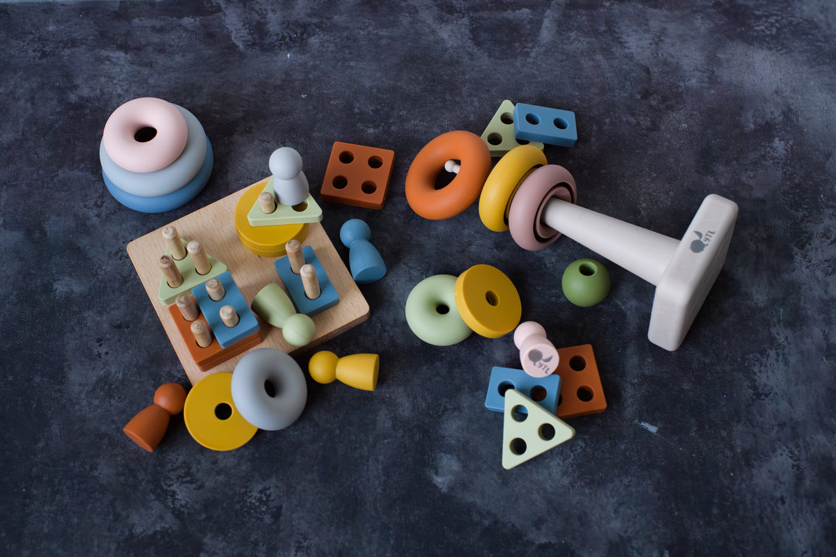 The Montessori Sensory Starter Kit – 3 Toy Set features wooden stacking rings, geometric shape sorters, and pegs scattered on a dark surface to support fine motor skills.