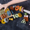A child’s hands playing with a Montessori Sensory Starter Kit – 3 Toy Set, featuring wooden geometric shapes and pegs in assorted colors to develop fine motor skills.