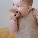 A baby wearing a floral headband chews on a wooden mushroom from the Mushroom Mix & Match Sorting Toy (24 pc), a toy designed to support fine motor skills.