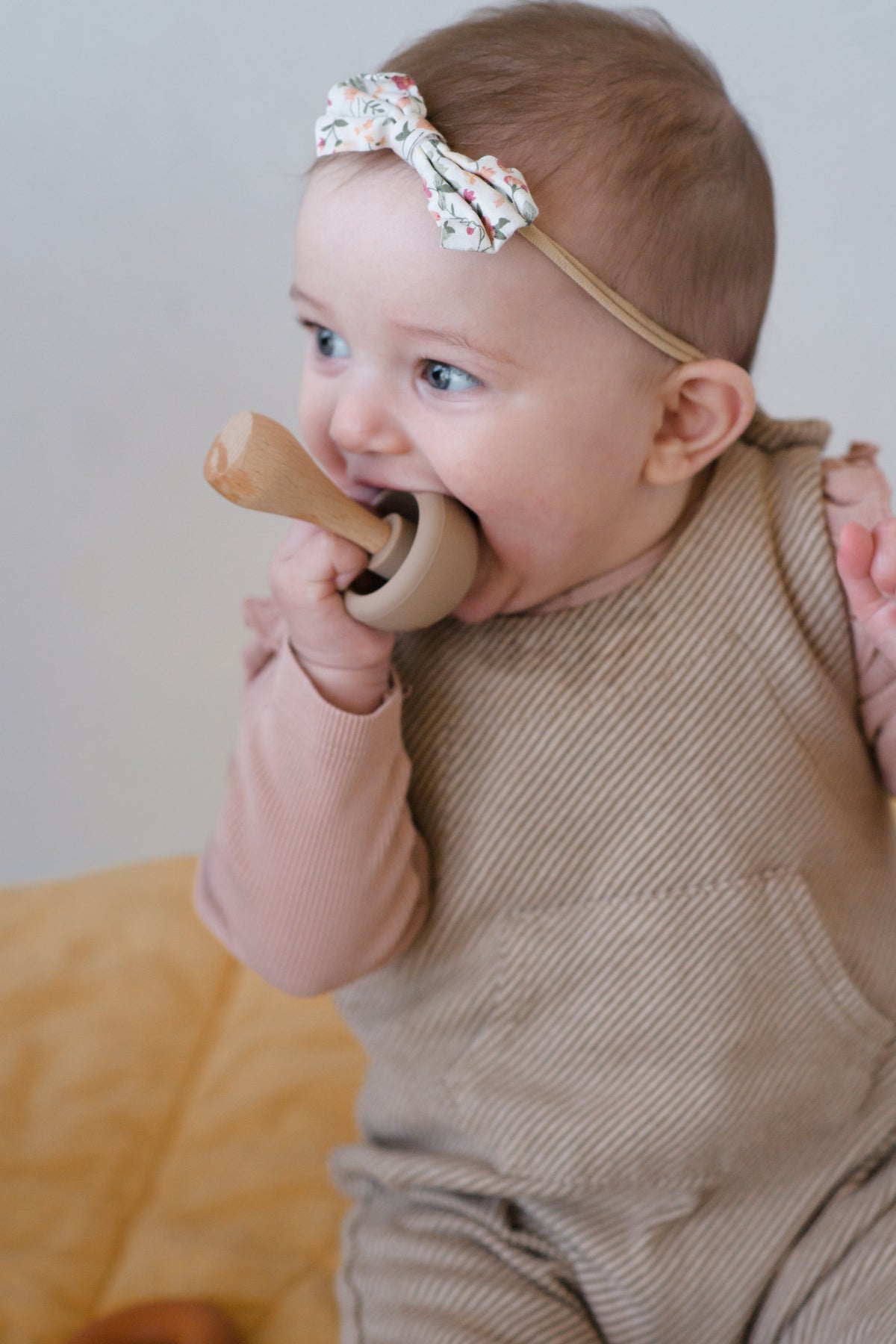 A baby wearing a floral headband chews on a wooden mushroom from the Mushroom Mix & Match Sorting Toy (24 pc), a toy designed to support fine motor skills.