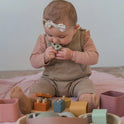 A baby sits on a pink blanket engaging in sensory play with the Montessori First Birthday Gift Set – 3 Toys, featuring wooden and silicone stacking pieces.