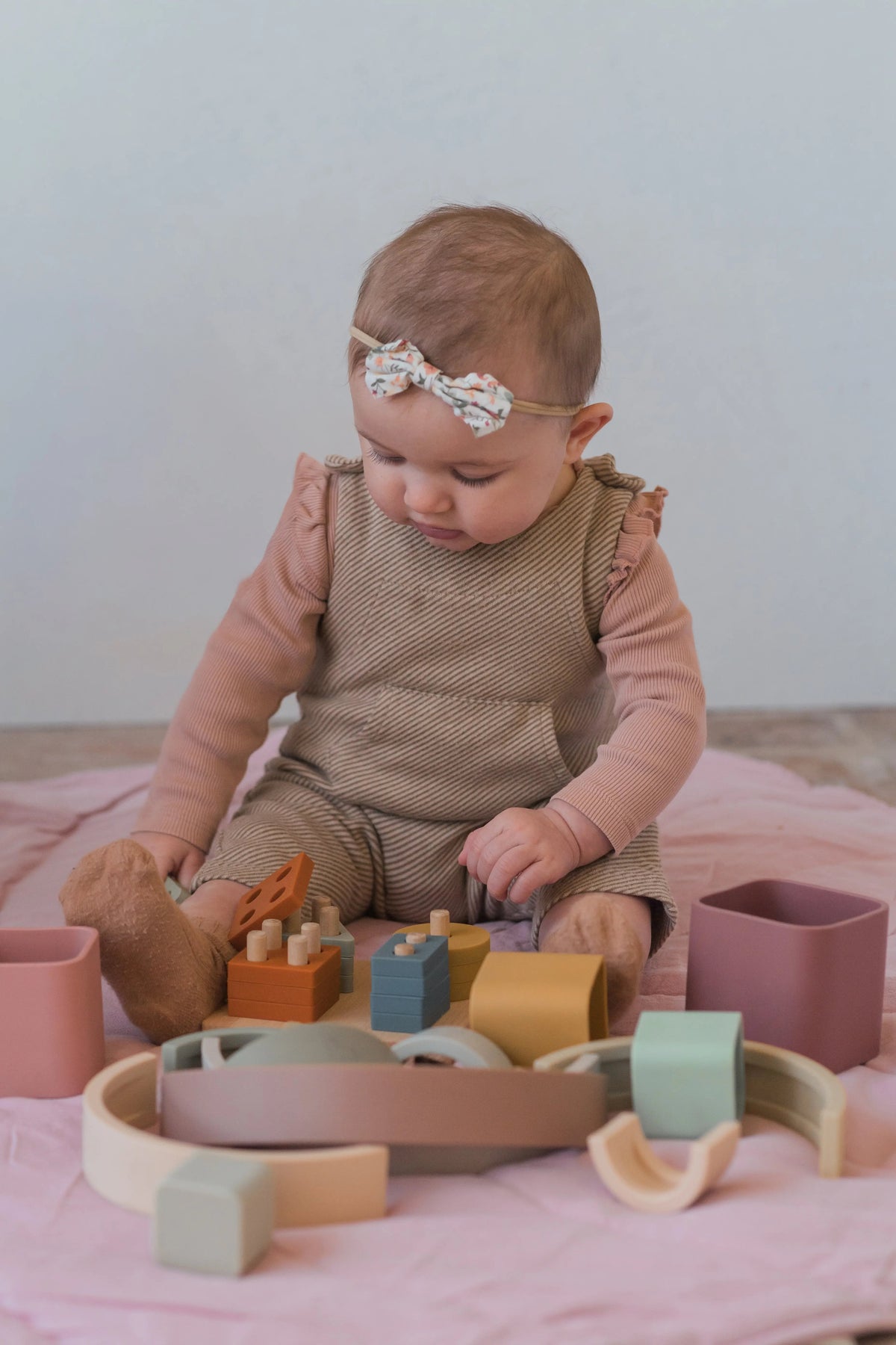 A baby sits on a pink mat engaging in sensory play with the Montessori First Birthday Gift Set – 3 Toys, featuring wooden and silicone stacking pieces.