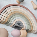 A 10-piece Silicone Rainbow Stacking Toy featuring muted rainbow arches surrounded by various small wooden and silicone play figures.