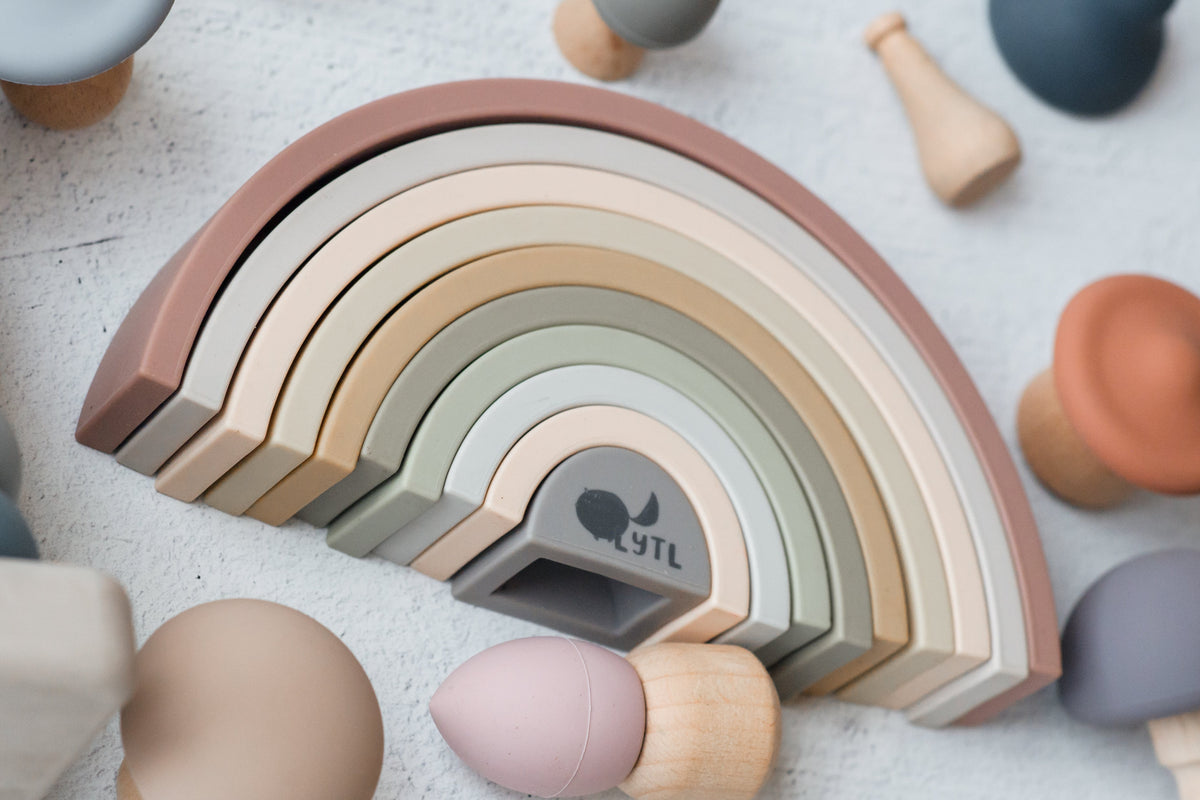 A 10-piece Silicone Rainbow Stacking Toy featuring muted rainbow arches surrounded by various small wooden and silicone play figures.