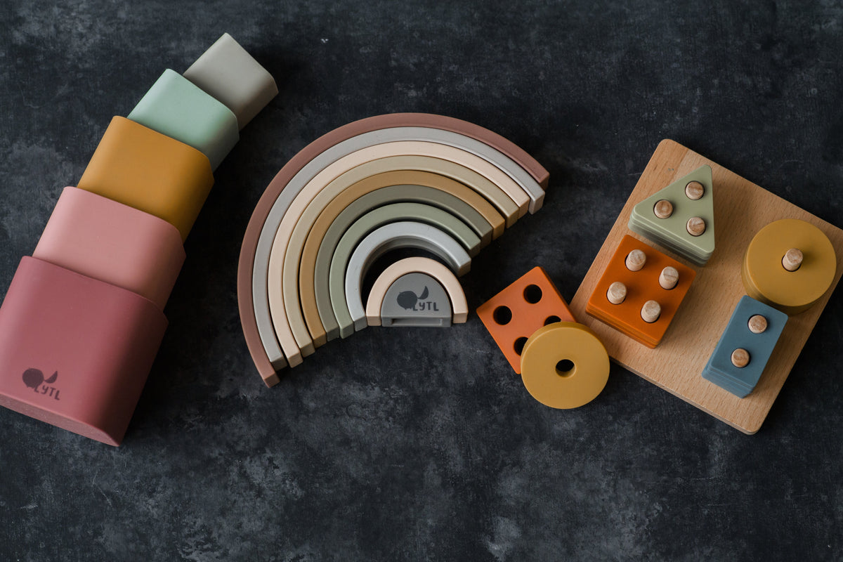 Silicone Stacking Cups (Set of 5), a nested rainbow-shaped stacking toy, and a wooden shape-sorting board with various blocks arranged on a dark surface.