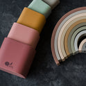 Five Silicone Stacking Cups (Set of 5) in a gradient of muted colors stacked next to a semicircular rainbow-shaped stacking toy.