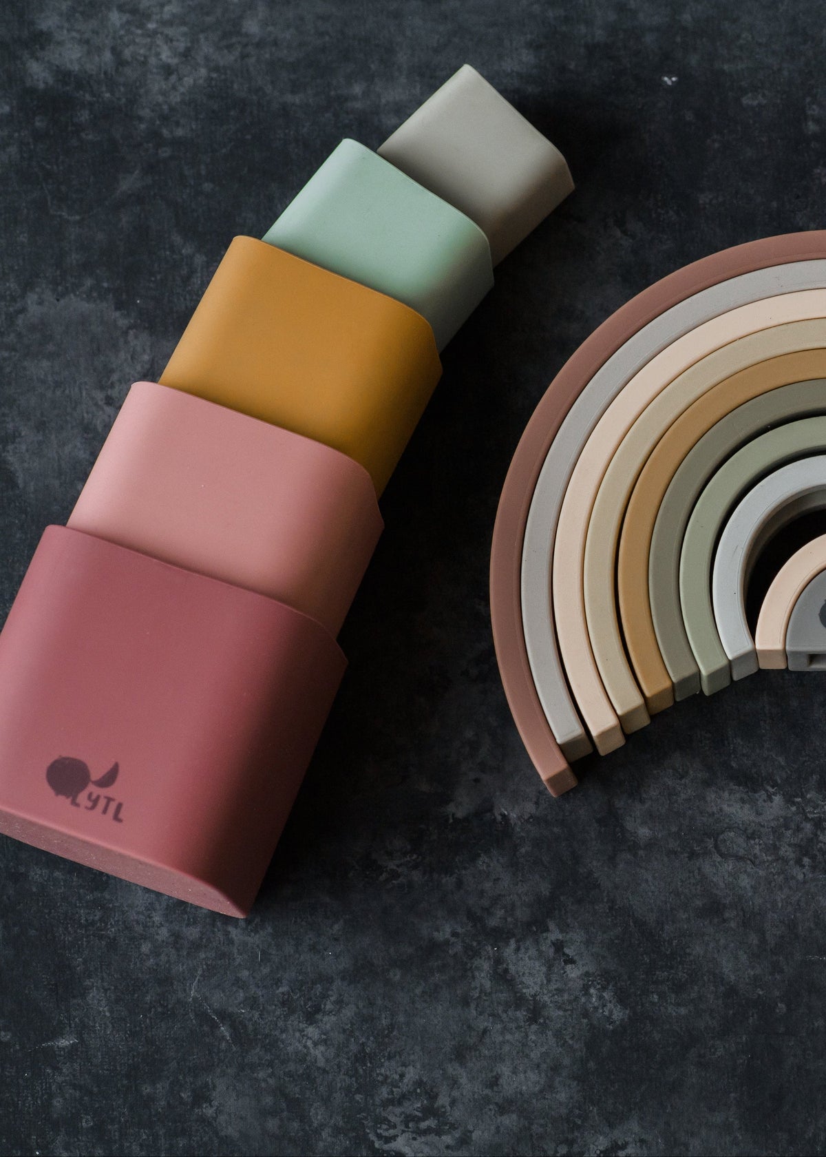 Five Silicone Stacking Cups (Set of 5) in a gradient of muted colors stacked next to a semicircular rainbow-shaped stacking toy.